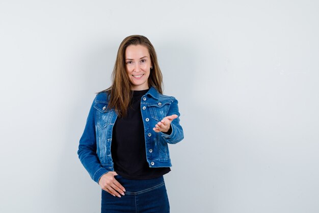 Young lady in blouse, jacket, jeans stretching hand to show something and looking confident