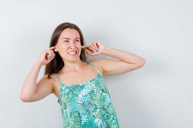 Young lady in blouse plugging ears with fingers and looking annoyed , front view.