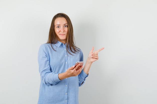 Young lady in blue shirt holding mobile phone while pointing away and looking curious