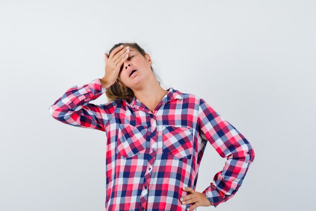 Young lady in checked shirt keeping hand on forehead and looking annoyed , front view.