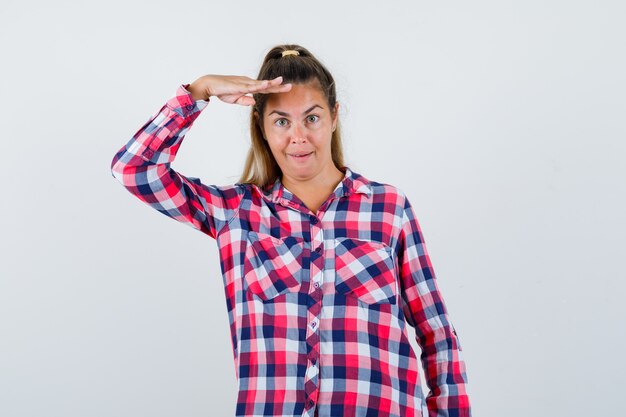 Young lady in checked shirt looking at camera with hand over head and looking astonished , front view.