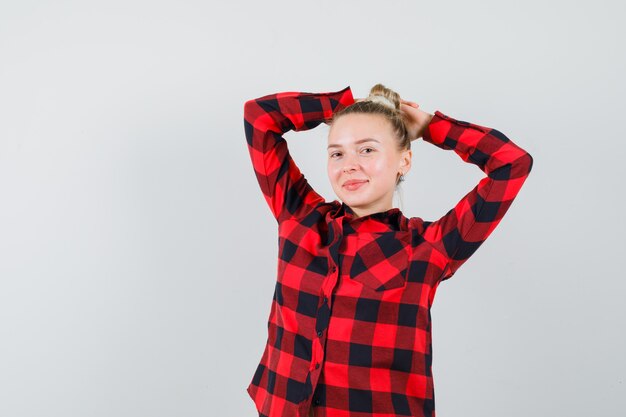 Young lady in checked shirt posing with raised hands behind head and looking glad