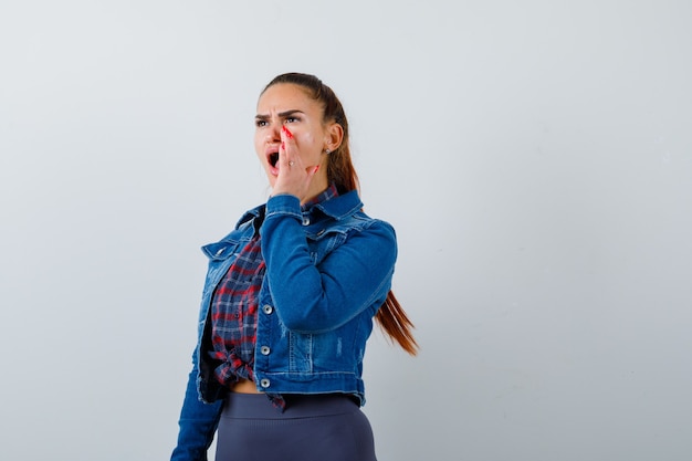 Free Photo young lady in checkered shirt, denim jacket shouting while with hand near mouth and looking annoyed , front view.