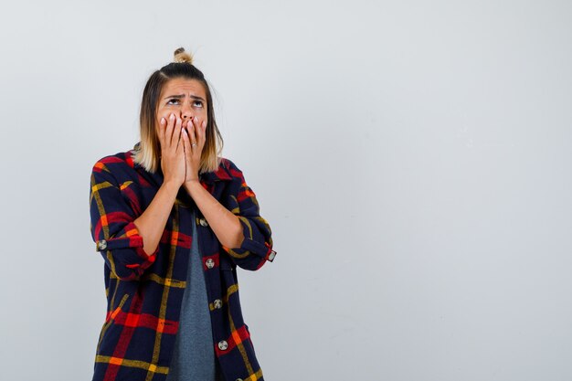 Young lady covering mouth with hands in casual checked shirt and looking upset , front view.