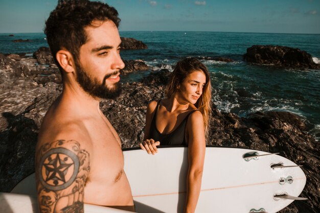 Young lady and guy with surf boards on rock shore near sea