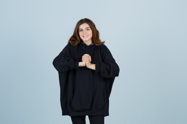 Young lady holding clasped hands in oversized hoodie, pants and looking hopeful , front view.