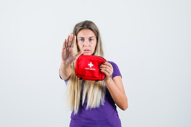 Young lady holding first aid kit, showing stop gesture in violet t-shirt , front view.