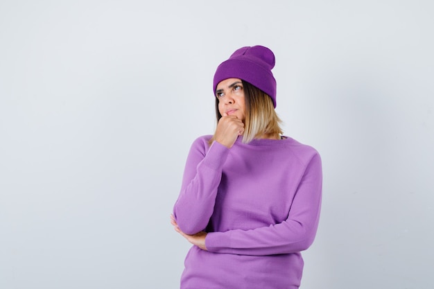 Young lady holding fist on chin in purple sweater, beanie and looking pensive. front view.