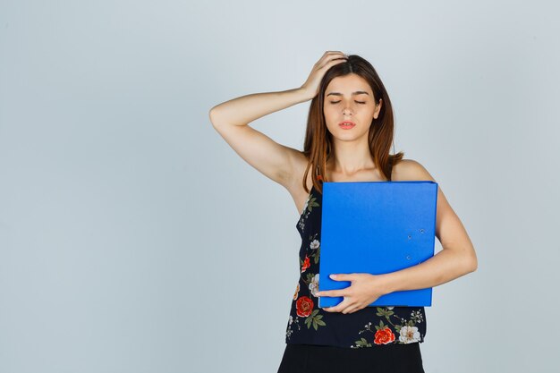 Young lady holding folder while keeping hand on head in blouse, skirt and looking tired , front view.