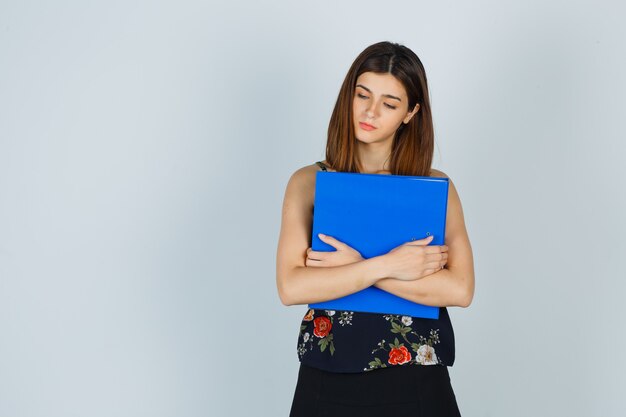 Young lady holding folder while looking down in blouse, skirt and looking gloomy. front view.