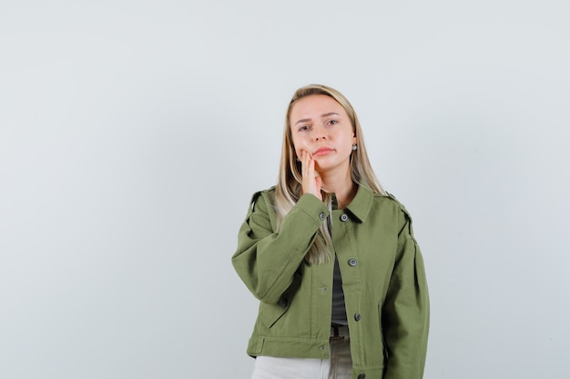 Young lady holding hand on cheek in jacket, pants and looking pensive. front view.