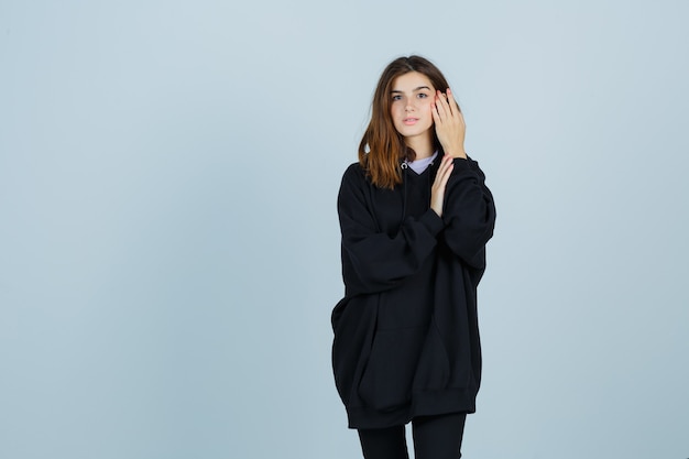 Young lady holding hand on cheek in oversized hoodie, pants and looking charming. front view.