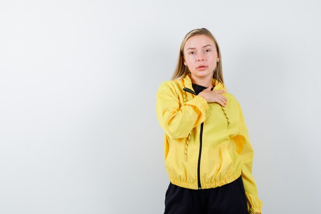 Young lady holding hand on chest in yellow jacket, pants and looking confident , front view.