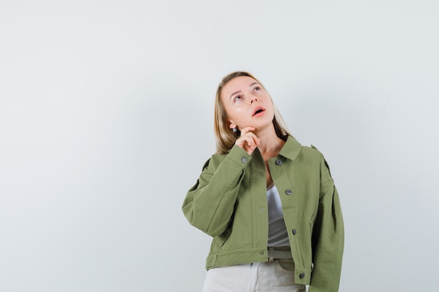 Young lady holding hand on chin in jacket, pants and looking pensive. front view.