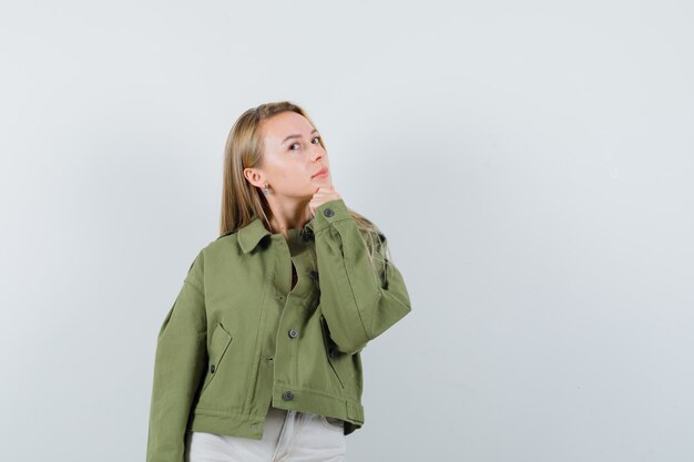 Young lady holding hand on chin in jacket, pants and looking pensive , front view.