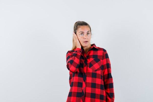 Young lady holding hand on ear in checked shirt and looking pensive