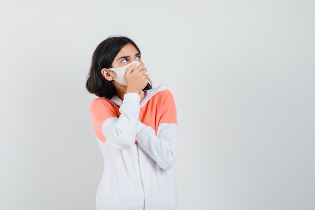 Young lady holding hand on her mouth in hoodie, face mask and looking scared.