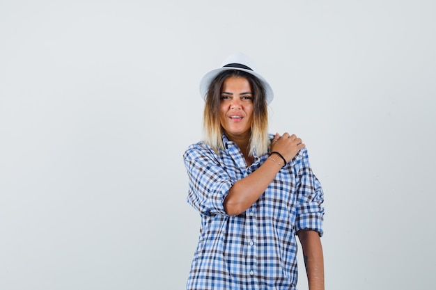 Young lady holding hand on shoulder in checked shirt hat and looking pretty 