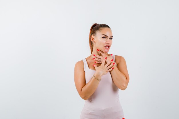 Young lady holding hand on throat and chest in beige tank top and looking confused