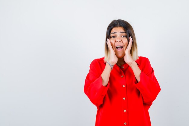 Young lady holding hands on cheeks in red oversize shirt and looking excited , front view.