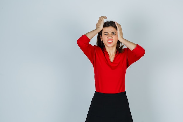 Free photo young lady holding hands on head in red blouse, skirt and looking exhausted