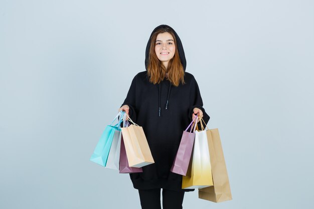 Young lady holding packets while looking at camera in oversized hoodie, pants and looking happy. front view.