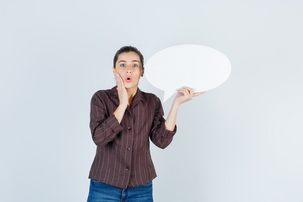 Young lady holding palm on cheek, keeping paper poster in shirt, jeans and looking surprised , front view.