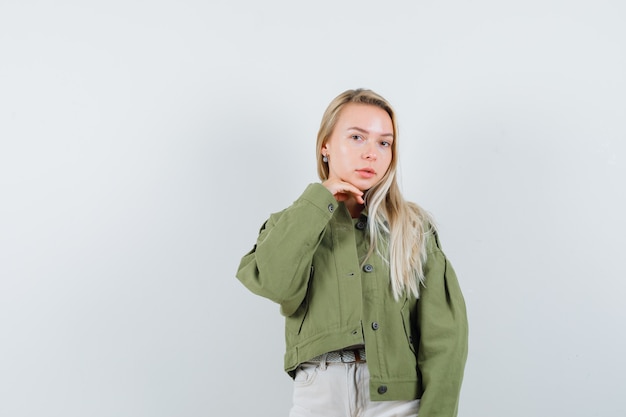 Young lady in jacket, pants holding hand under chin and looking cute , front view.