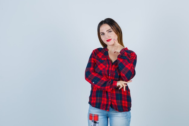 Young lady keeping finger on cheek in checked shirt, jeans and looking confident. front view.