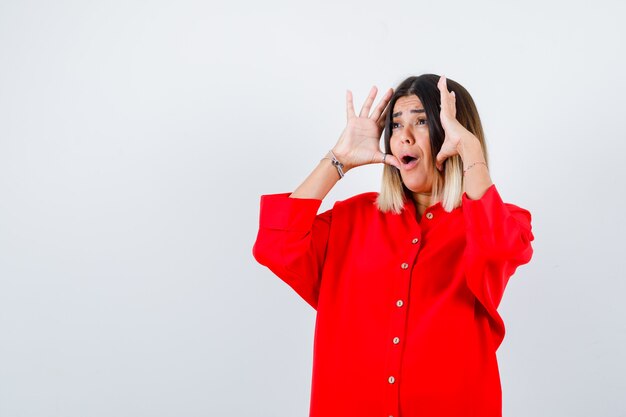 Young lady keeping hands near the face in red oversize shirt and looking scared front view.