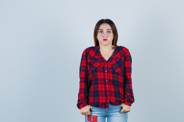 Young lady keeping thumbs in pockets while blowing cheeks in checked shirt, jeans and looking bored , front view.