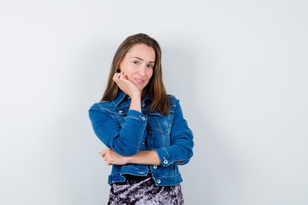 Young lady leaning cheek on hand in denim jacket and looking sensible. front view.