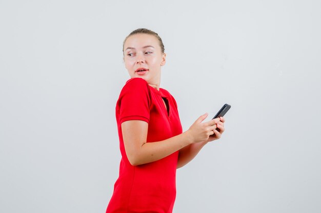 Young lady looking back while holding mobile phone in red t-shirt and looking curious .