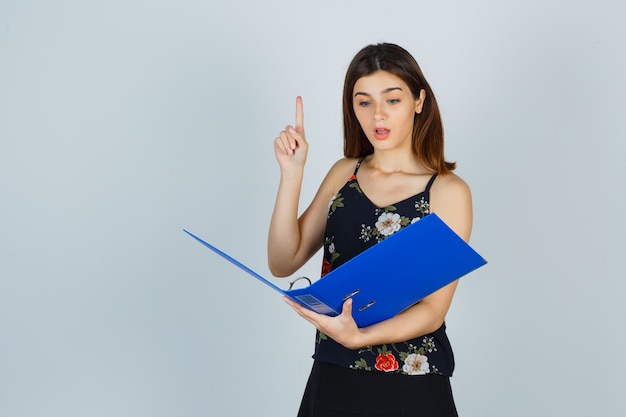 Young lady looking over documents in folder, showing eureka gesture by pointing up in blouse, skirt and looking wondered , front view.