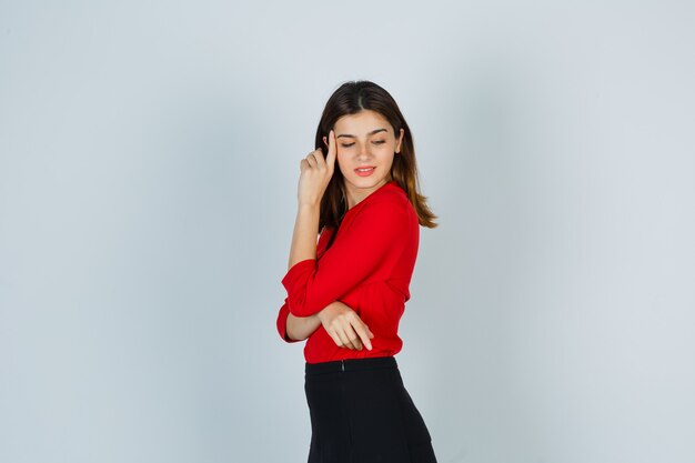 Young lady looking down while posing in red blouse, skirt and looking delicate