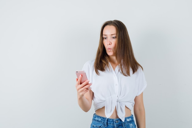 Young lady looking at phone in white blouse and looking surprised. front view.