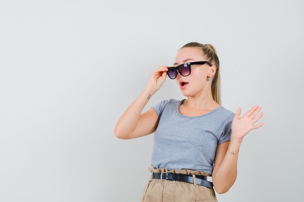 Young lady looking through glasses and waving hand in t-shirt and pants