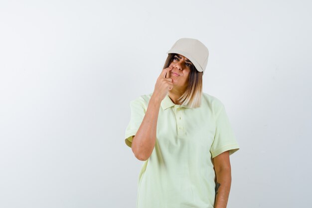 Young lady looking up while thinking in t-shirt, cap and looking thoughtful. front view.