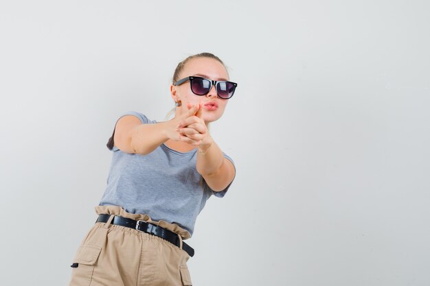 Young lady making finger pistol sign in t-shirt and pants and looking confident