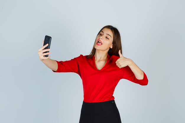 Young lady making video call while showing thumb up in red blouse