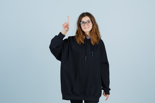 Young lady in oversized hoodie, pants pointing up and down and looking happy , front view.