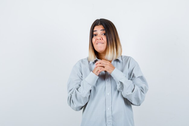 Young lady in oversized shirt holding clasped hands over chest and looking anxious , front view.