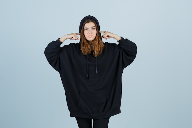 Free photo young lady plugging ears with fingers in oversized hoodie, pants and looking annoyed , front view.