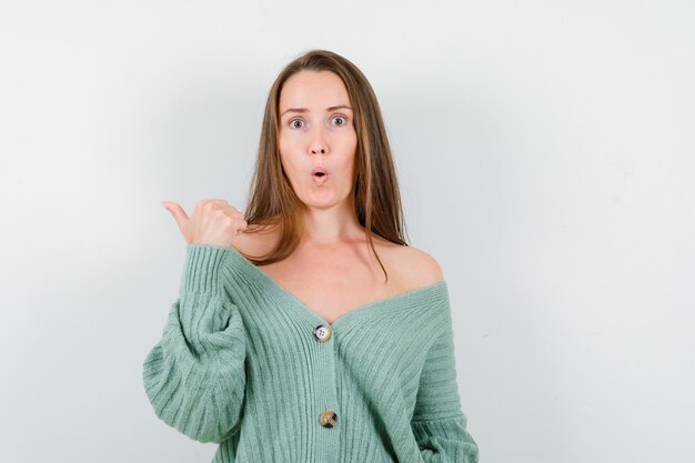 Young lady pointing aside with thumb in wool cardigan and looking surprised. front view.