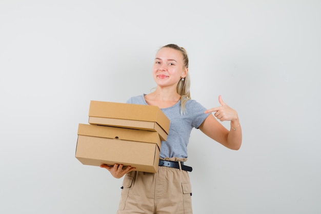 Young lady pointing at cardboard boxes in t-shirt and pants and looking merry