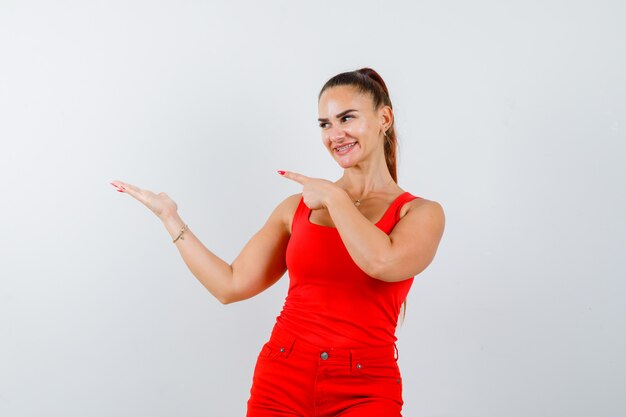 Young lady pointing to the left side in red singlet, red trousers and looking merry , front view.