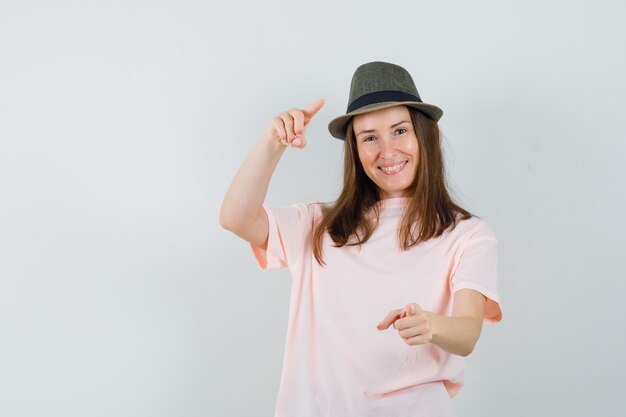 Young lady pointing in pink t-shirt, hat and looking happy , front view.