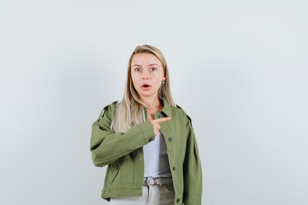 Young lady pointing to the right side in jacket, singlet and looking surprised. front view.