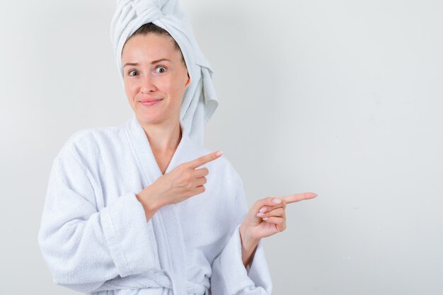 Young lady pointing to the right side in white bathrobe, towel and looking funny. front view.