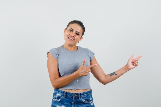 Young lady pointing to the side in t-shirt, shorts and looking cheerful. 